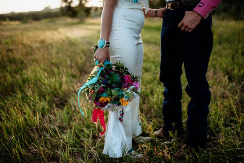 Bride and groom stand facing holding bouquet at side in grassy field 