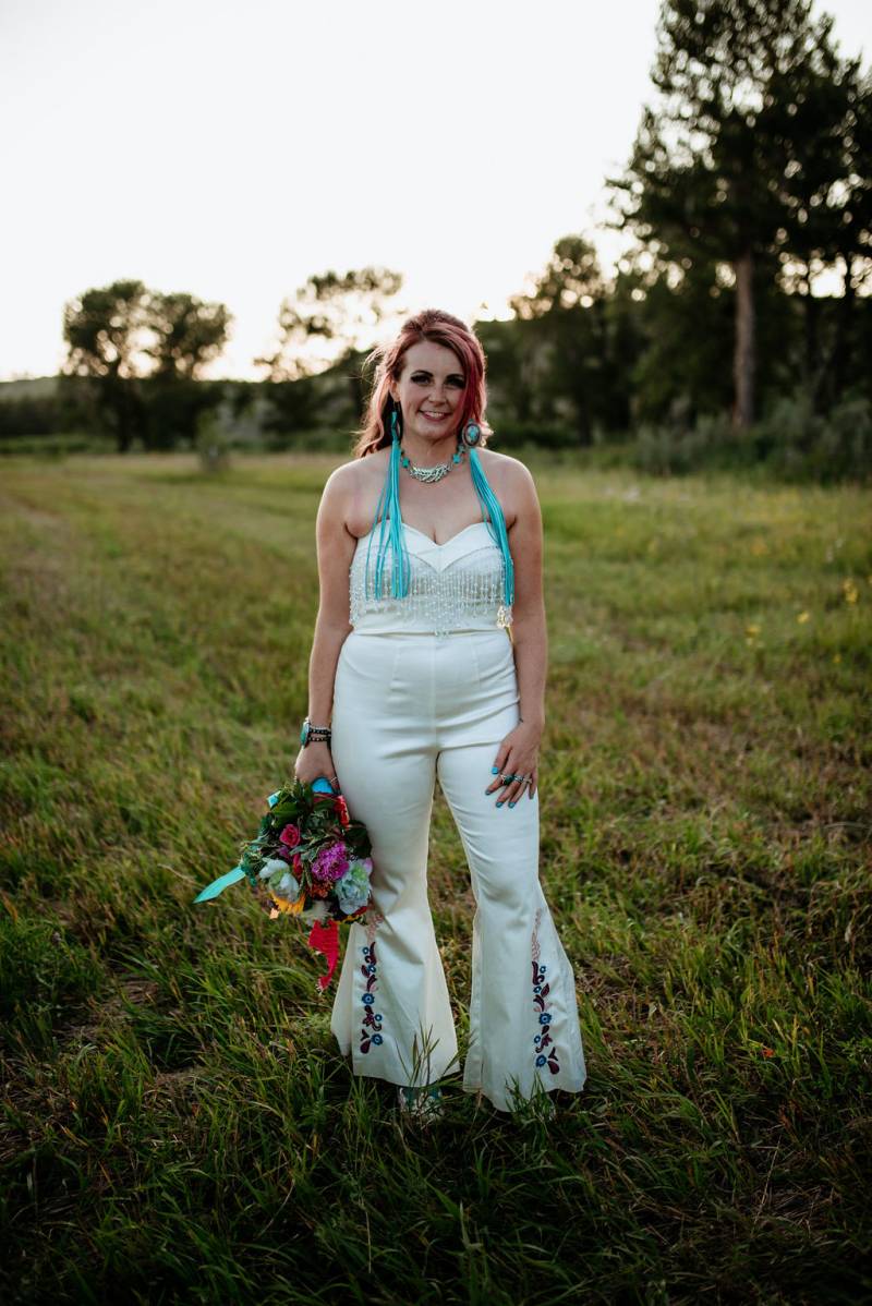 Bride stands in white dress holding bouquet to side in grassy field 