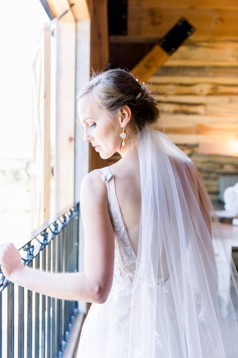 Bride stands against window looking over shoulder in white dress and veil