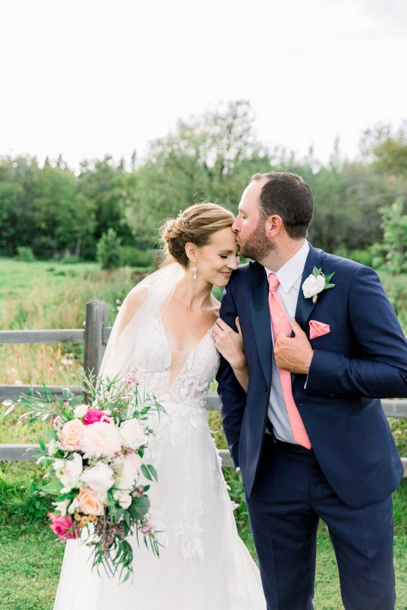 Groom kisses brides forehead while holding white and pink bouquet 