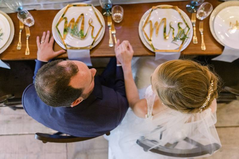 Bride and groom sit at table with gold cutlery holding hand 