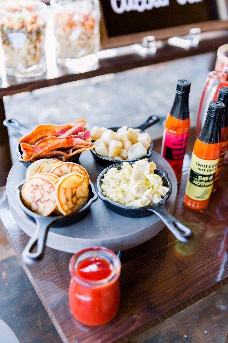 Mini cast-iron pans with breakfast foods beside hot sauces on brown table 