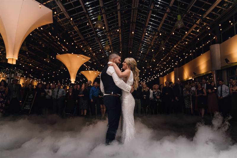 Man and woman embrace in large auditorium lit with fairy lights while fog rolls beneath them  