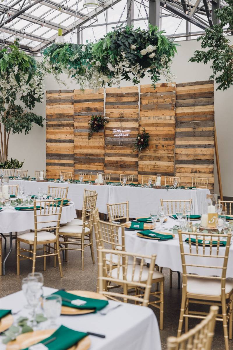 Large wood backboard behind head table with large green potted trees and hanging white flower accents 