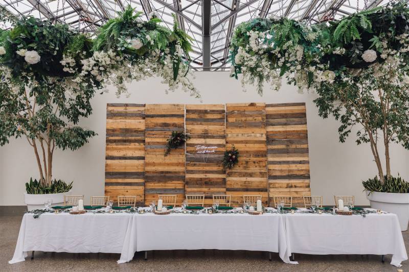 Large wood backboard behind head table with large green potted trees and white flower accents 