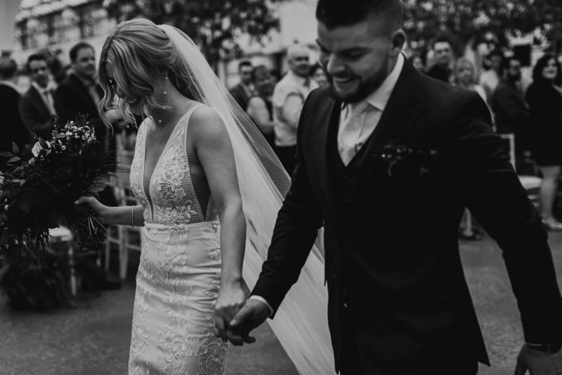 Woman in long white veil and white dress walk holding hands with man in dark suit 