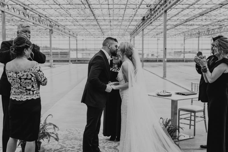 Man and woman kiss while family applauds to the side in large glass venue