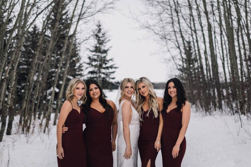 Five women in white and burgundy dresses standing in snowy forest pathway 