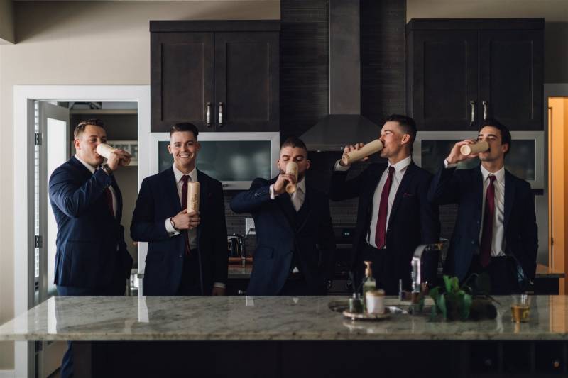 Five men in navy suits drinking from tall beige cups in kitchen