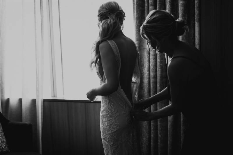 Women zipping up white lace open back dress on another woman beside window