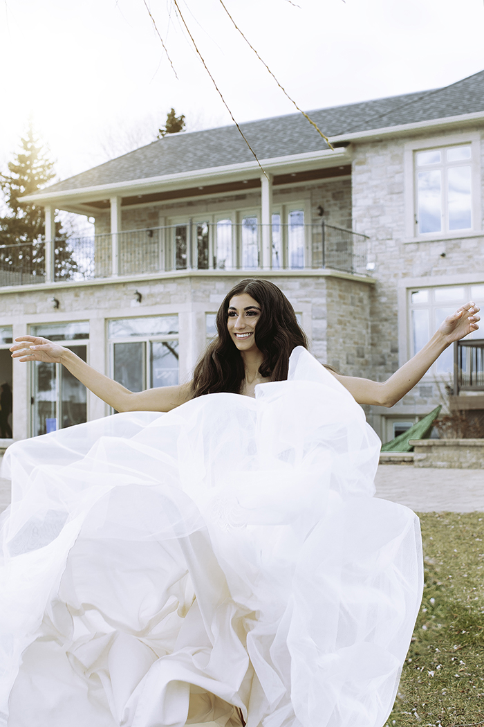 Woman throws up skirt smiling hands to side in front of large house 