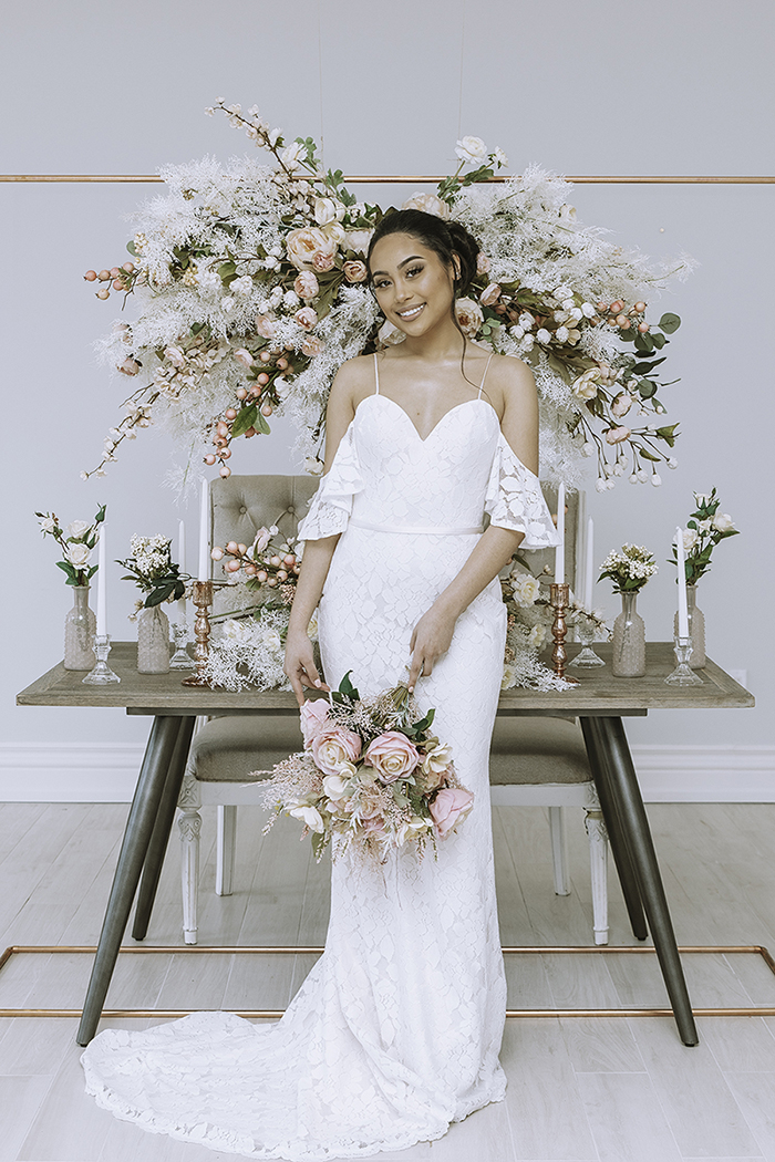 Woman in white dress standing in front of hanging floral arrangement over table with large blush centerpiece and candlesticks