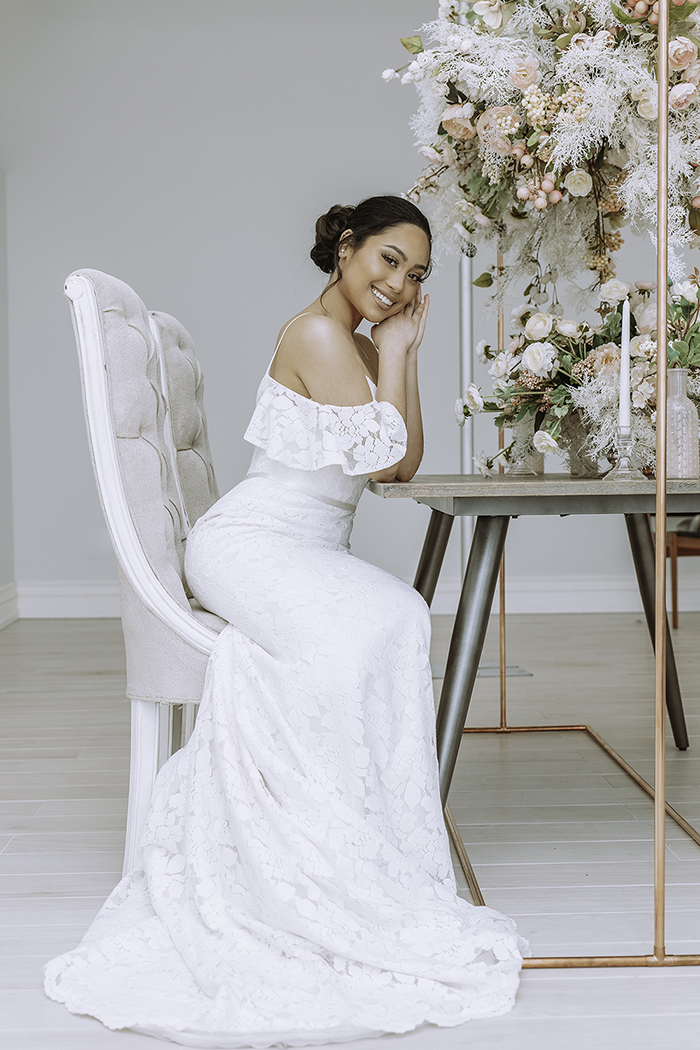Woman in white dress sits leaning against hands by table with large blush floral arrangement 