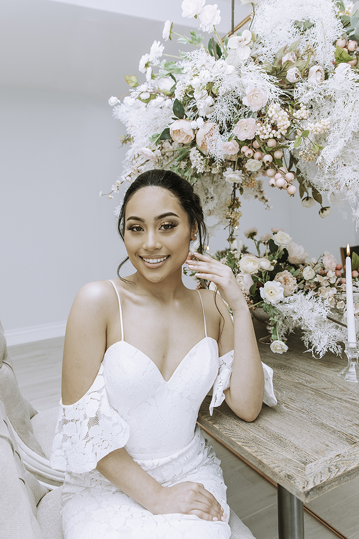 Woman sitting in white shoulder less dress leaning on table with large blush floral arrangement behind 