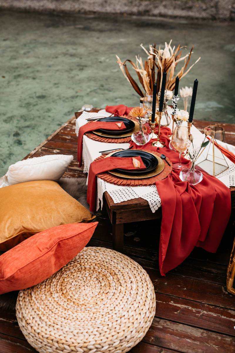 Small table on dock with orange and yellow pillows macramé and red fabric table runner and place settings 
