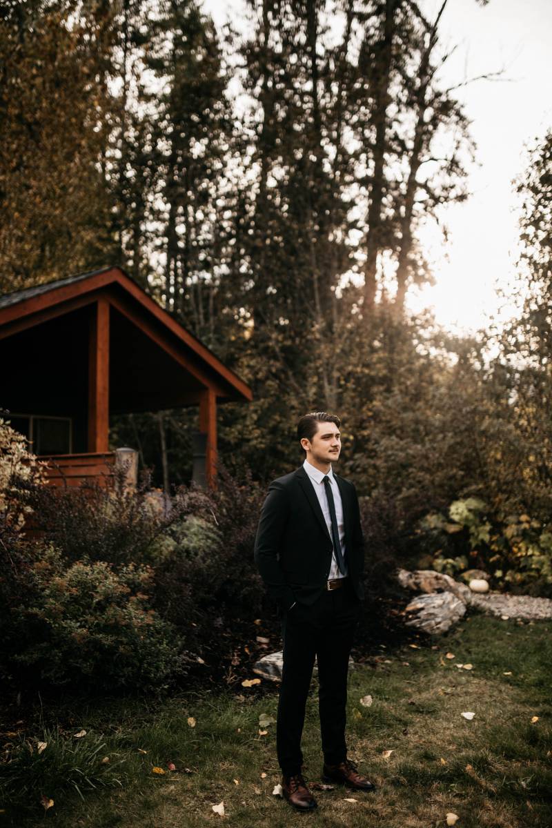 Man in suit stands in grassy wooded area in front of dark red wood cabin