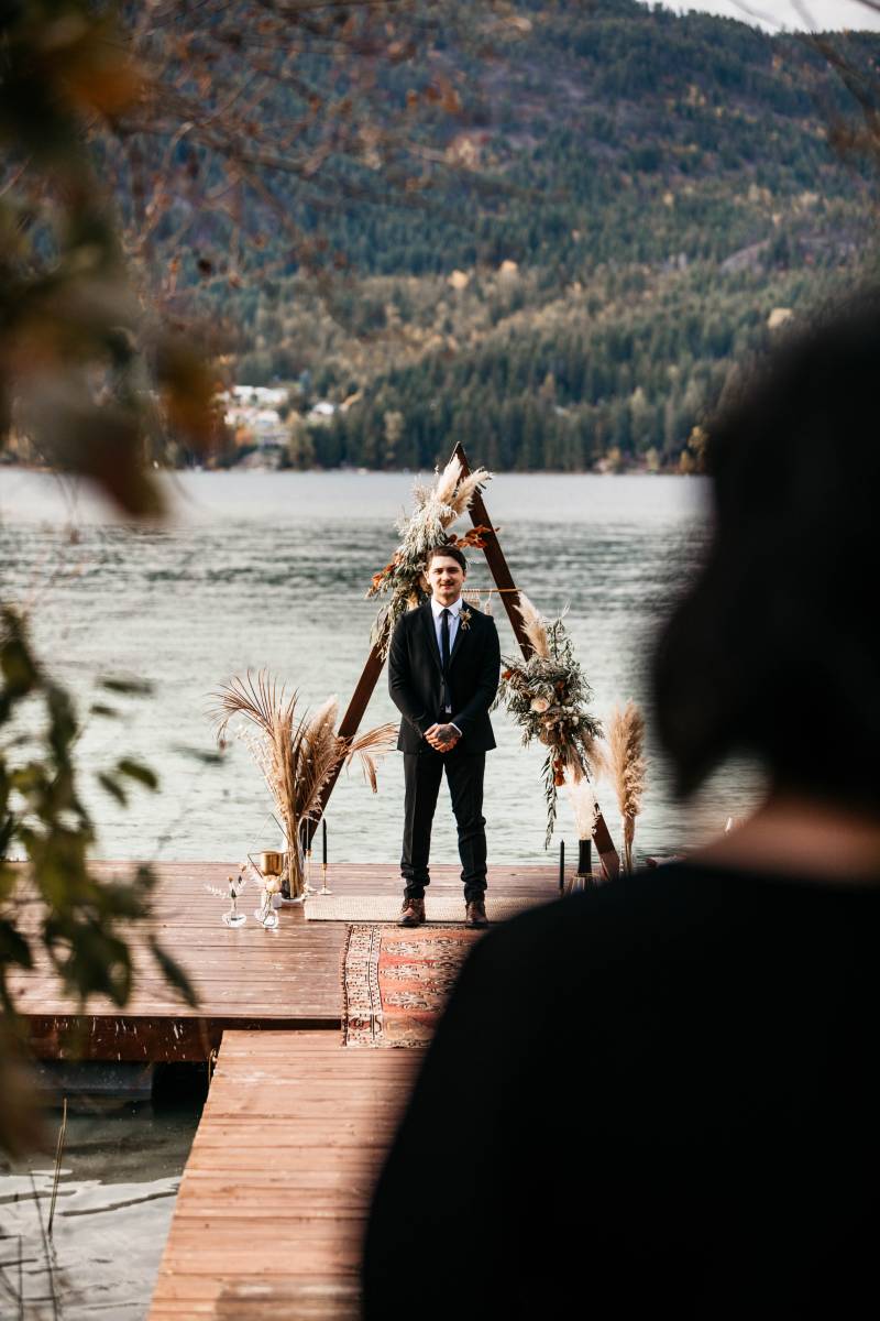 Man in suit standing in front of triangular wedding arch with pampas grass accents at the end of dock facing lake