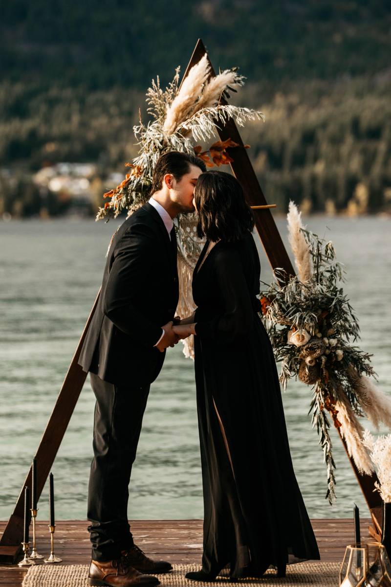Man in black suit and woman in black dress kiss in front of triangular wedding arch on dock 