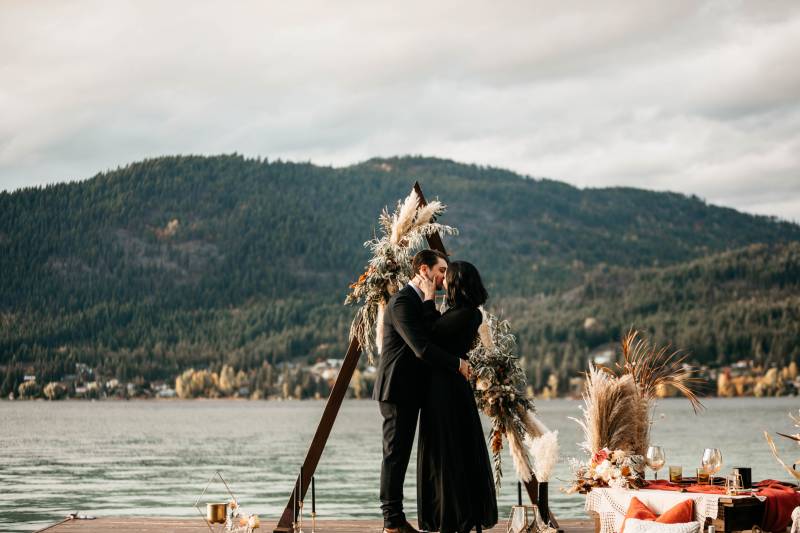 Man in black suit and woman in black dress embrace and kiss in front of triangular wedding arch on dock 