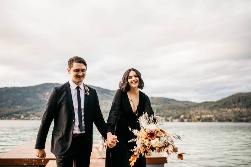 Man and woman walking down dock holding hands and holding white orange and peach bouquet