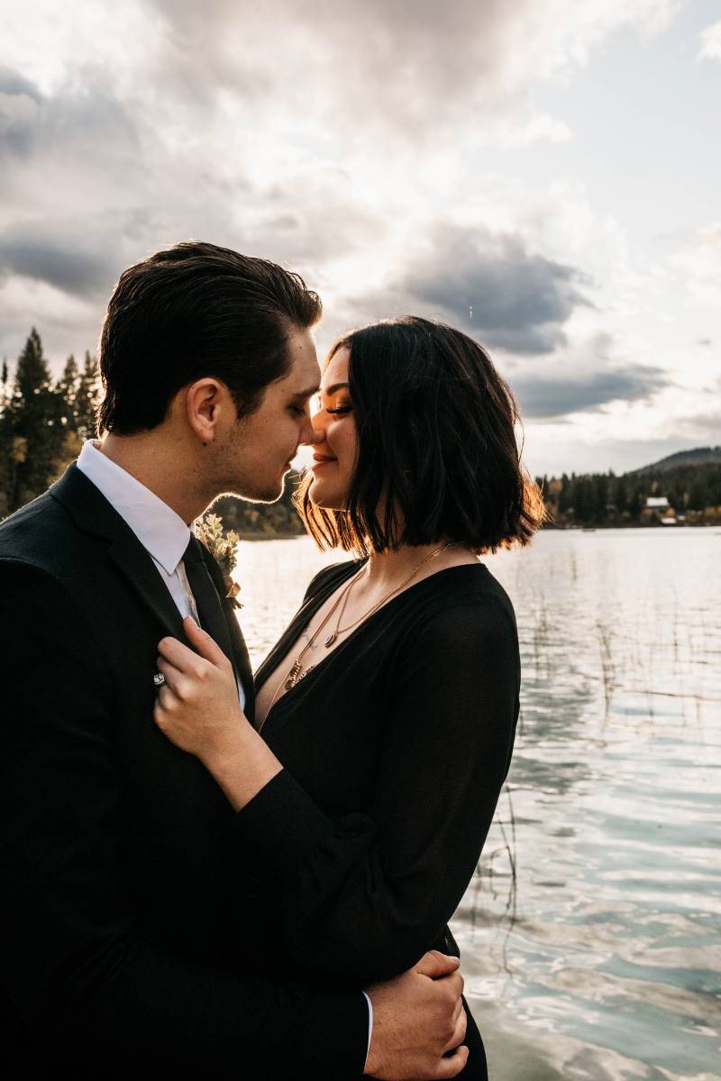 Man and woman in black suit and black dress embrace by edge of lake 