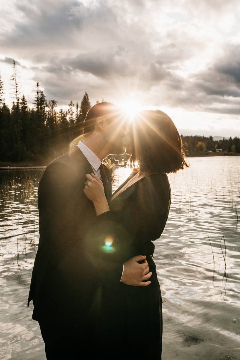 Man and woman embrace and kiss in front of lake with lens flare 