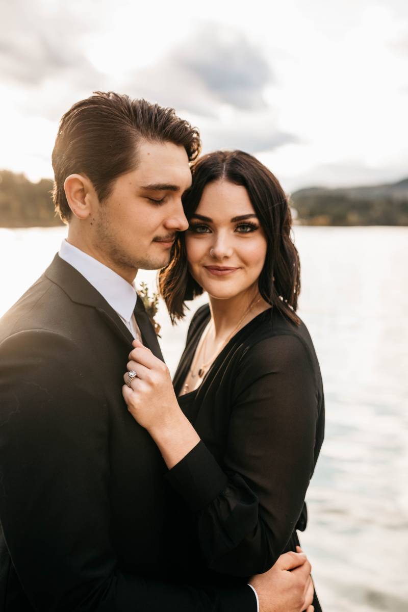 Man and woman in black suit and black dress embrace in front of lake 