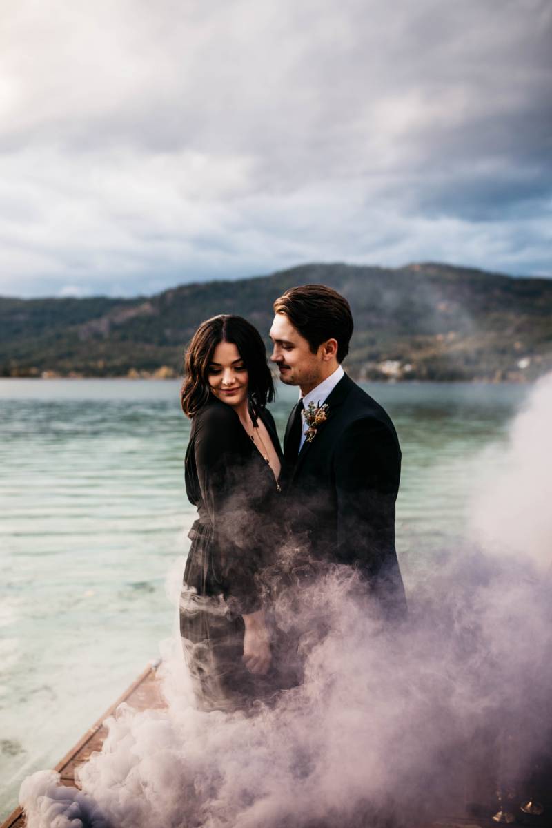 man and woman in black embrace while smoke wraps around them in front of lake