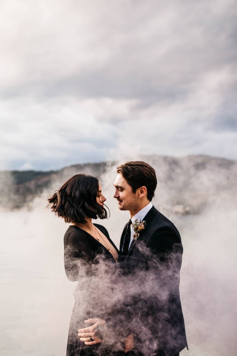 man and woman in black embrace while smoke wraps around them 