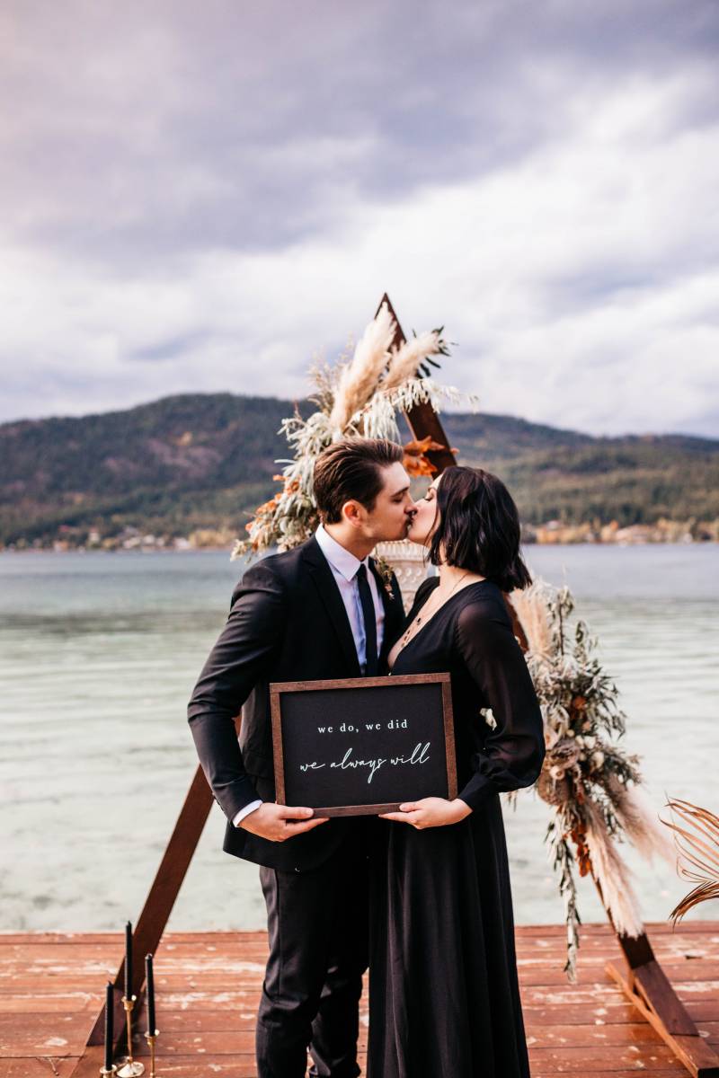 Man and woman kiss holding black sign on dock in front of triangular wedding arch