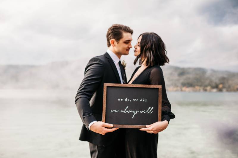 Man and woman embracing while holding wood framed sign in front of lake 