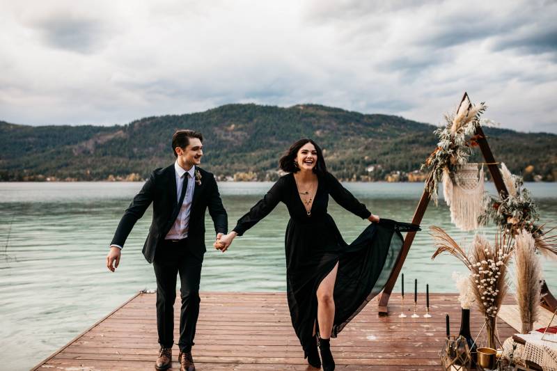 Man and woman with flowing black dress holding hands walking down dock in front of triangular wedding arch and pampas grass
