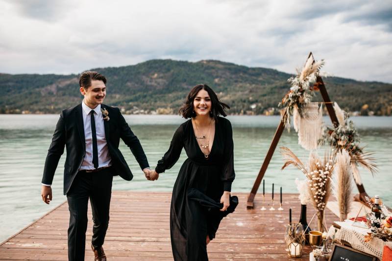 Man and woman holding hands walking down dock in front of triangular wedding arch and pampas grass
