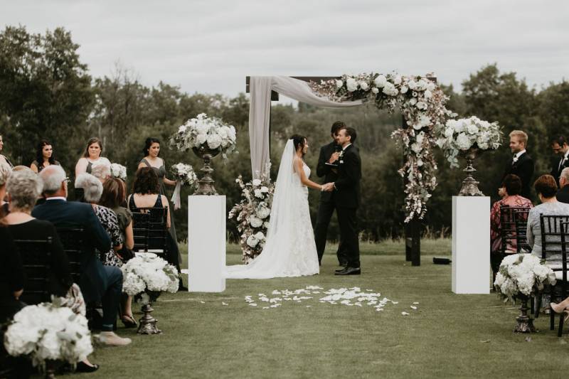 Bride and groom stand holding hands under wedding arch with white pedals on ground 