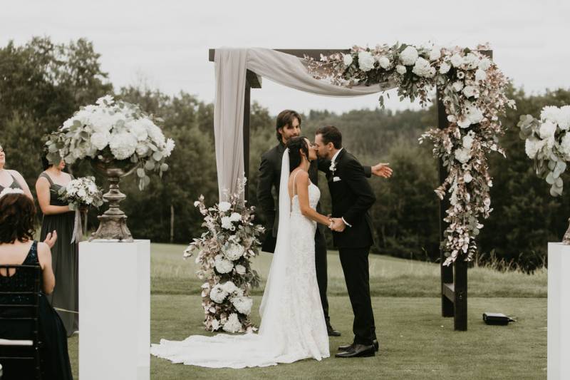 Bride and groom kiss underneath wedding arch with white flowers and fabric  