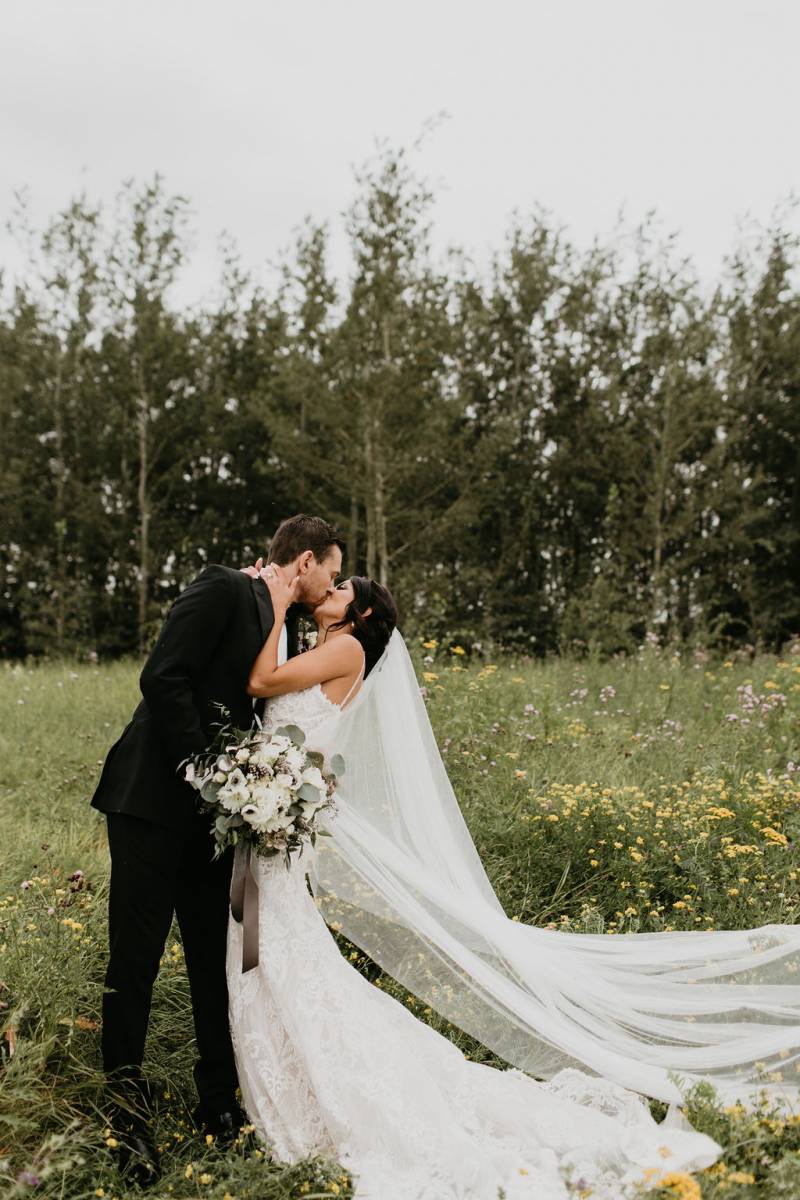 Bride and groom kiss while veil flows in grassy field and groom holds white bouquet 