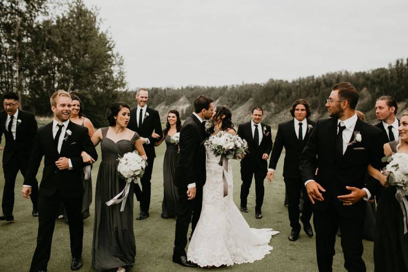 Bride and groom kiss while walking between bridesmaids and groomsmen in field 