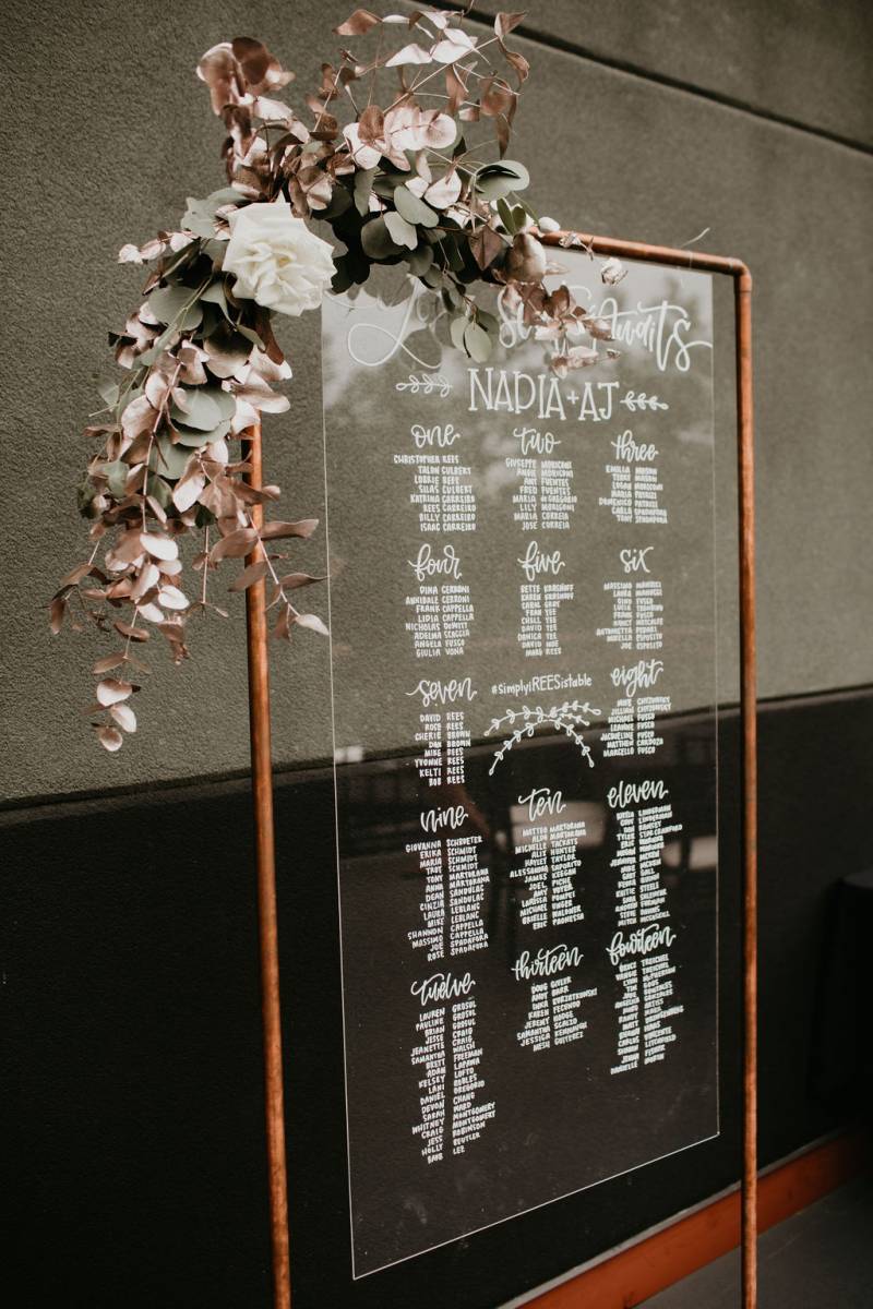 Wedding seating chart on clear pane hanging from bronze tube with blush floral accent 