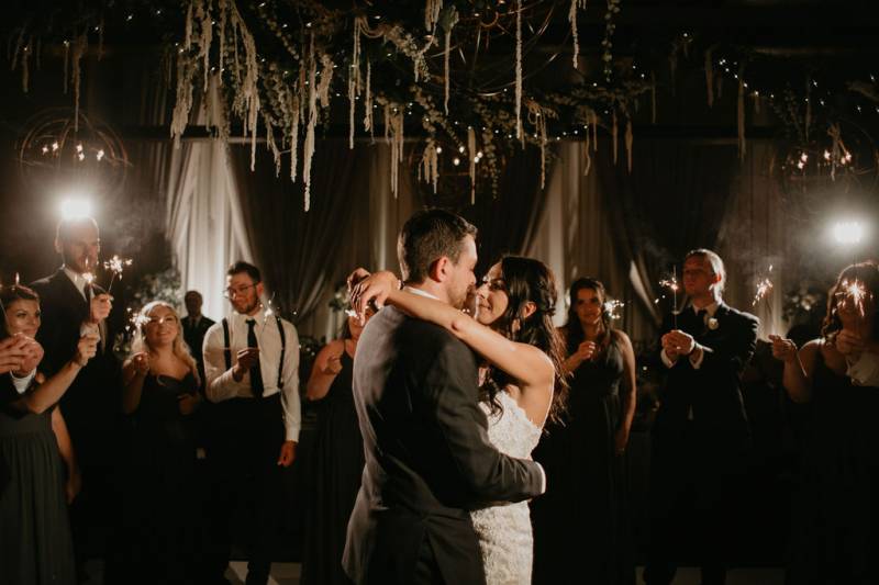 Bride and groom embrace while guests hold sparklers in dim room 