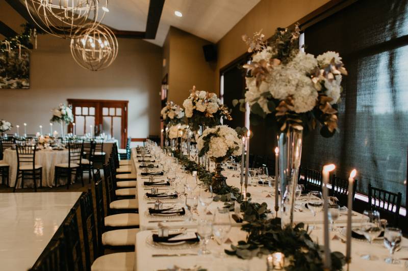 Long white tables set with candles and long green table runner and large white floral centerpieces 