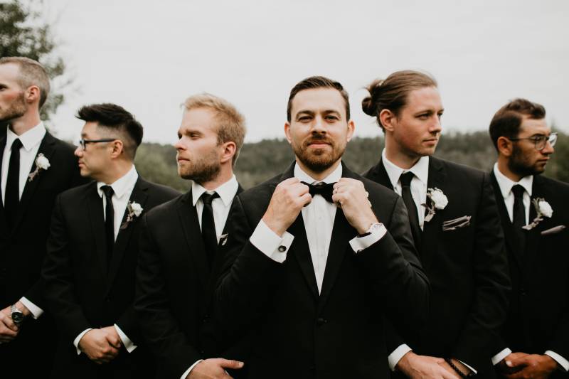 Groom and groomsmen standing together in black suits while groom adjusts bowtie 