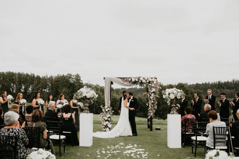 Bride and groom stand holding hands under wedding arch with white pedals on ground 