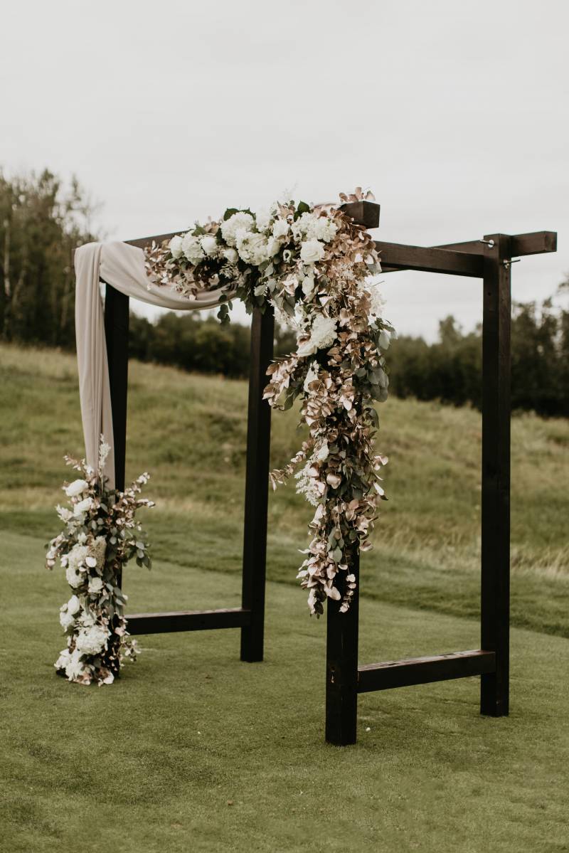 Large black wooden wedding arch with hanging white and beige flowers and hanging fabric in field 