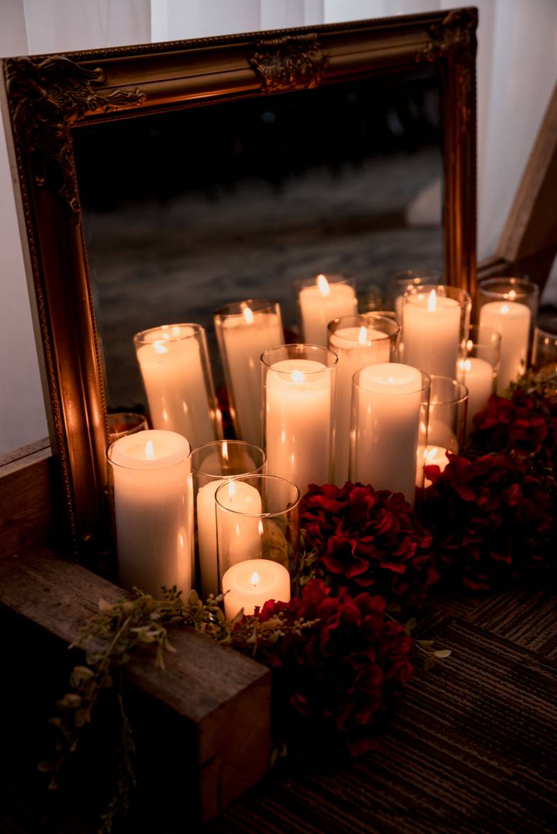 Several candles lit with burgundy floral arrangement around it in front of mirror 