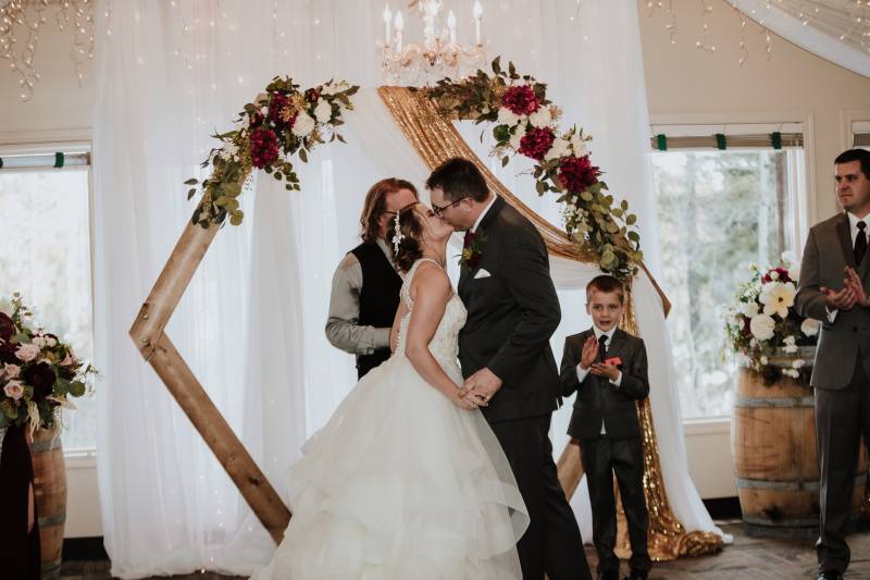 Bride and groom kiss in front of wooden wedding arch with gold and burgundy accents 