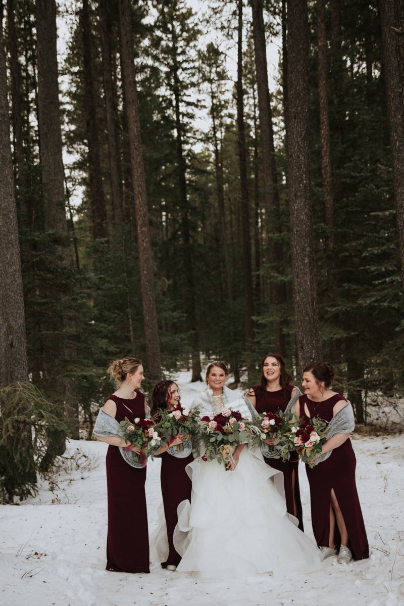 Bride and bridesmaids stand together holding blush and burgundy bouquets on snowy forest pathway 