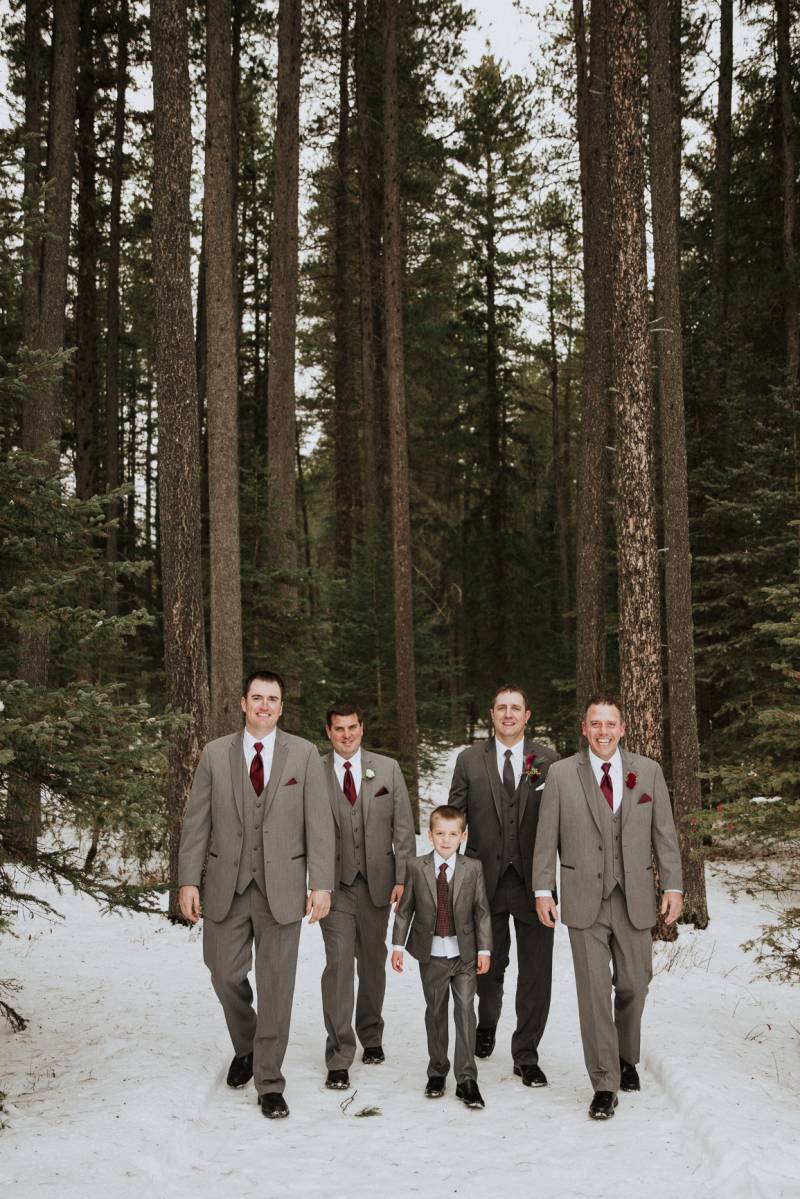 Groom and groomsmen and child walk together in front of tall trees on snowy pathway 