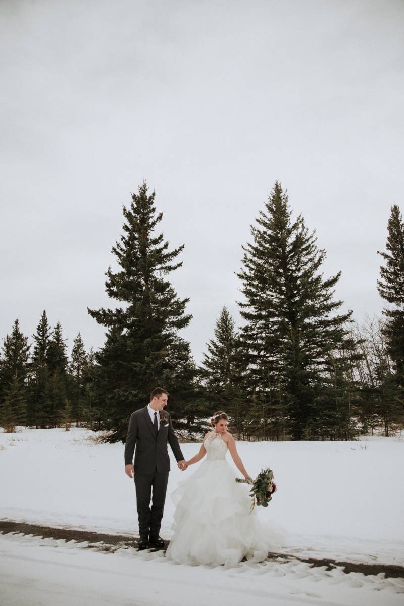 Bride and groom holding hands on snowy pathway holding bouquet out to side 
