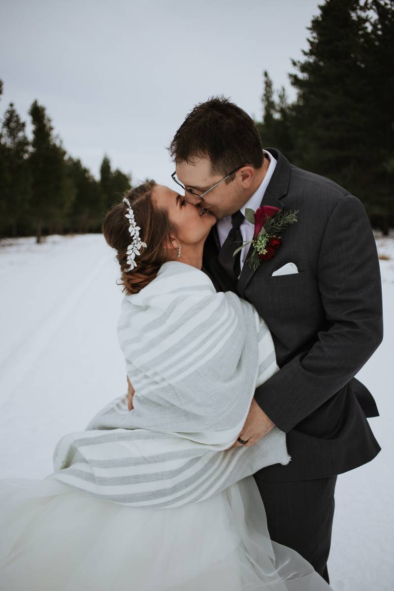Bride and groom kiss standing on snowy road in front of dense forest 