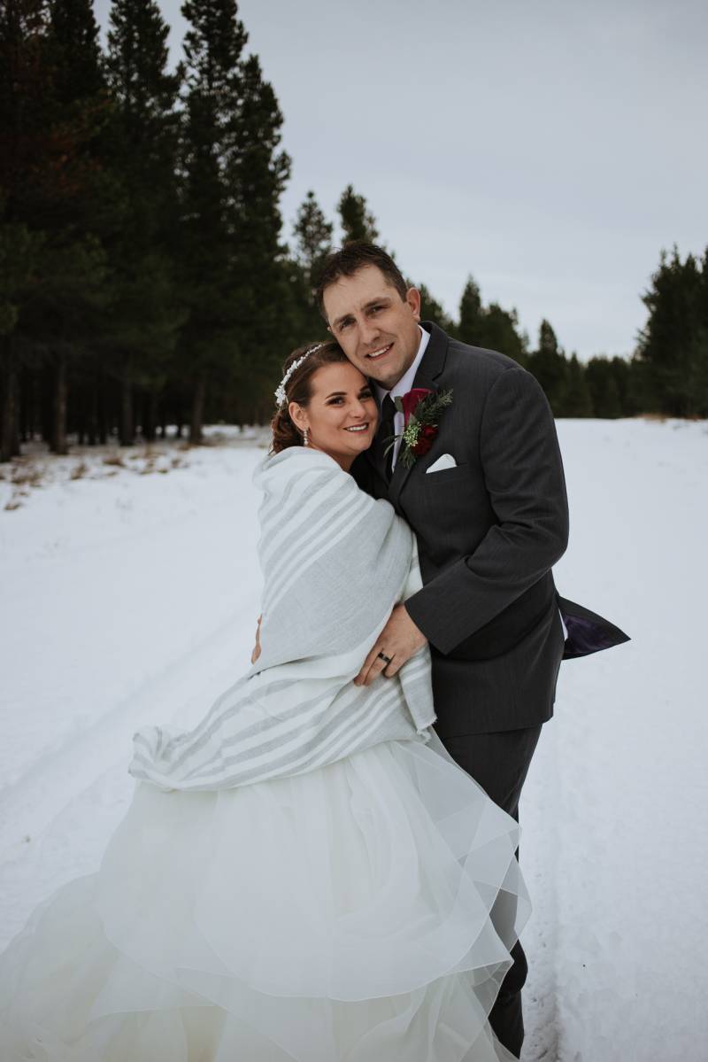 Bride and groom embrace on snowy pathway in front of dense green forest
